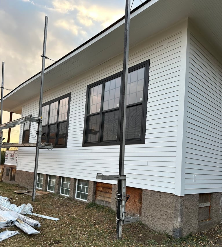 A white house under construction with newly installed black-framed windows on the upper floor. Scaffolding is set up in front of the house, and the basement level has small windows with stone and wood paneling. The word